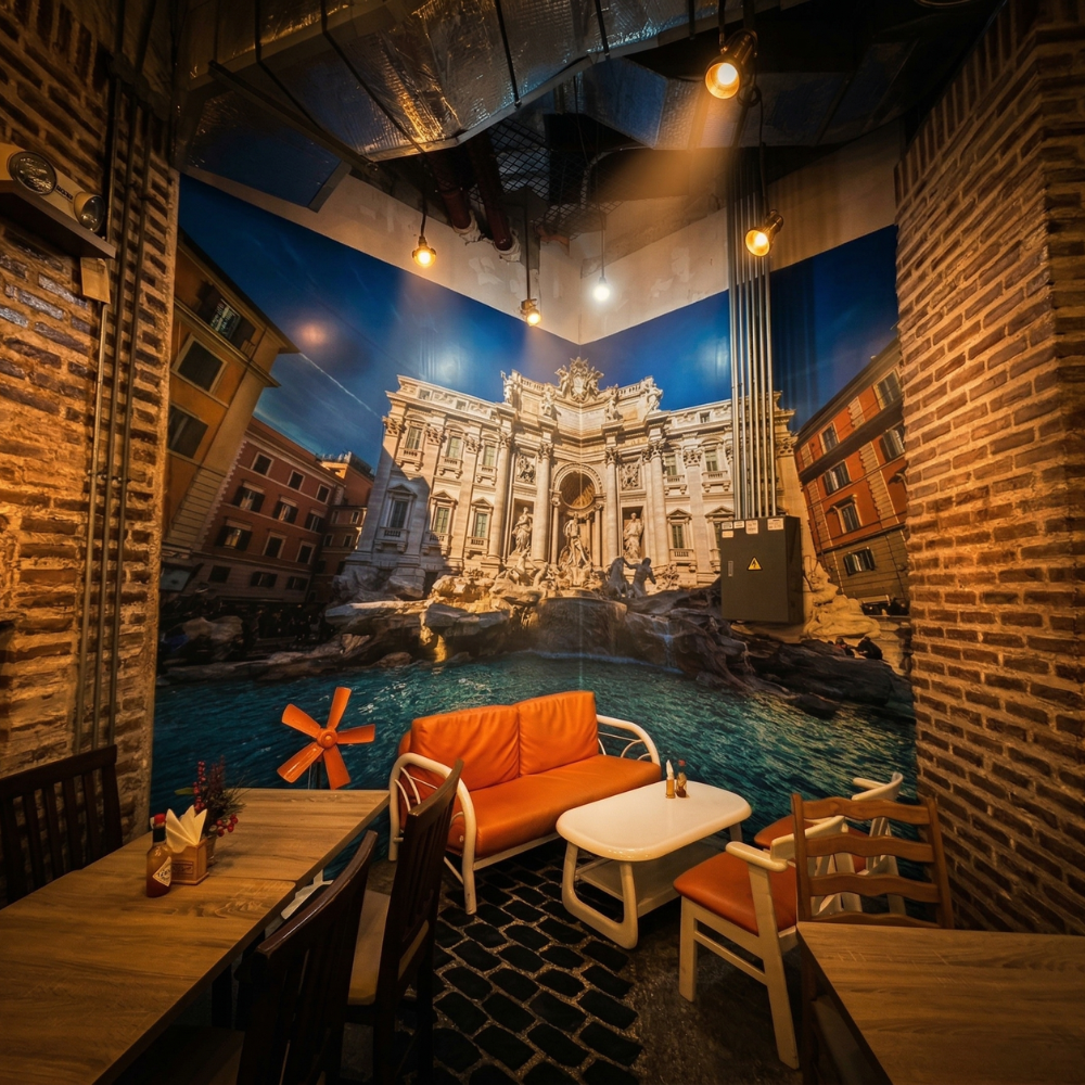 A large mural of Trevi Fountain covers the wall behind orange seating around wooden tables.