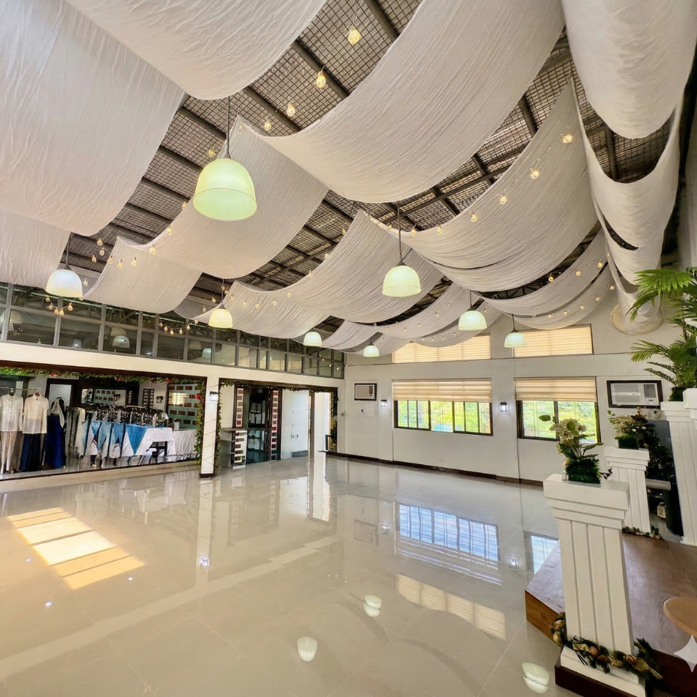 A spacious room with glossy floors, draped white fabric on the ceiling, hanging lights, large windows, and neatly arranged plants.