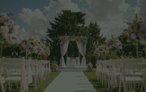 A beautiful outdoor wedding venue setup on a sunny day, showcasing white folding chairs with pink ribbons and a floral canopy in front of large green trees.