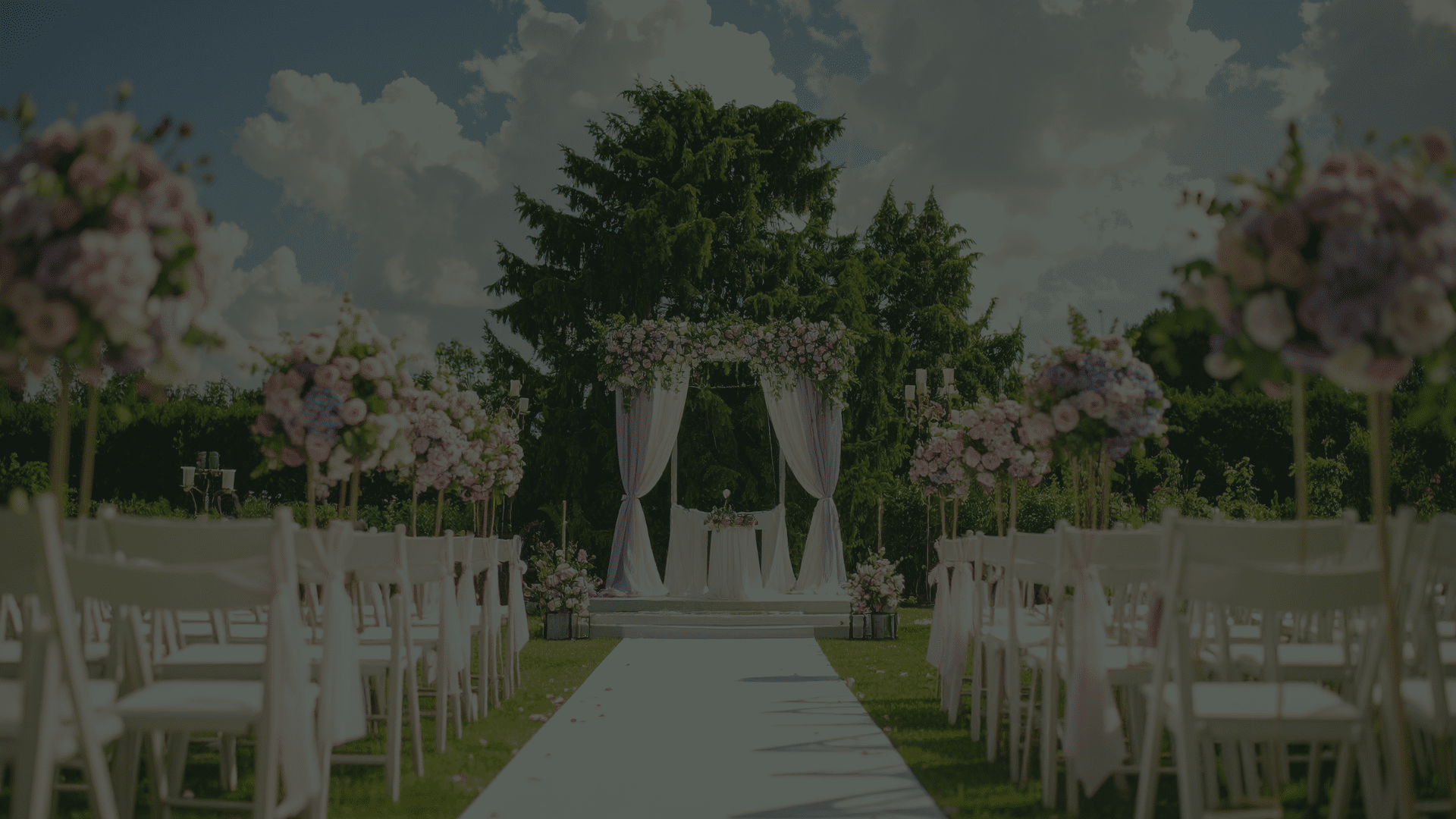A beautiful outdoor wedding venue setup on a sunny day, showcasing white folding chairs with pink ribbons and a floral canopy in front of large green trees.