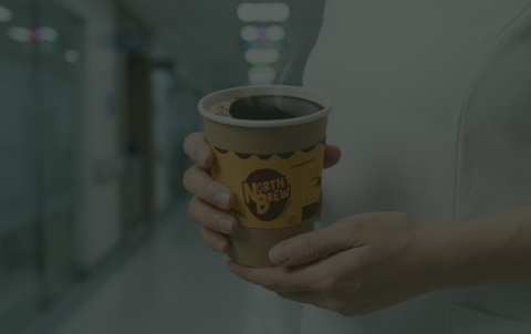 A close-up of a healthcare worker in a white uniform holding a steaming cup of black coffee in a North Brew Coffee branded paper cup, standing in a blurred hospital hallway.