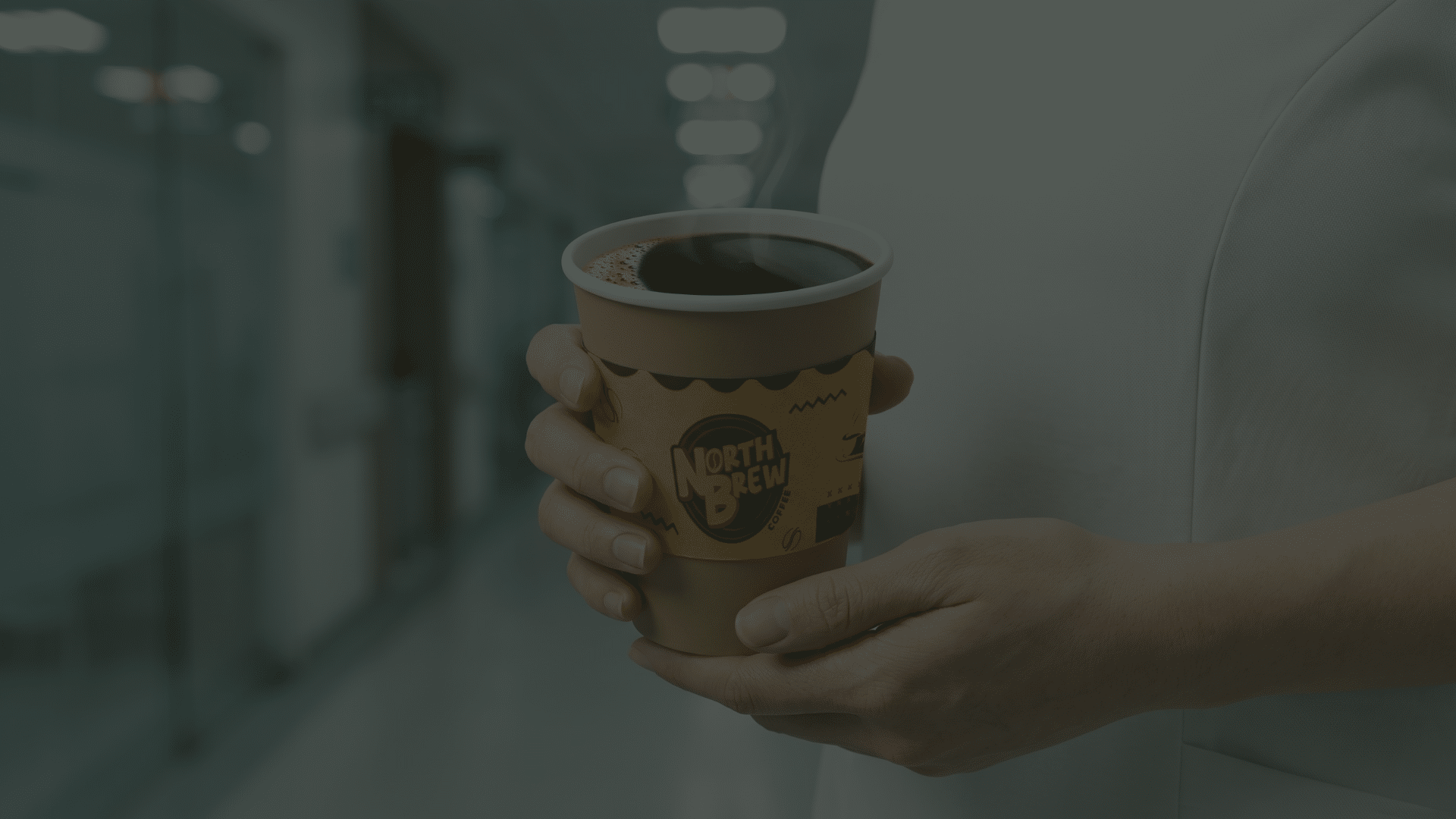 A close-up of a healthcare worker in a white uniform holding a steaming cup of black coffee in a North Brew Coffee branded paper cup, standing in a blurred hospital hallway.