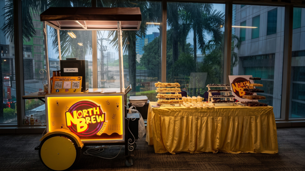 A North Brew Coffee mobile catering setup featuring an illuminated yellow coffee cart with an espresso machine next to a yellow-draped table filled with tiered pastries for an indoor event.