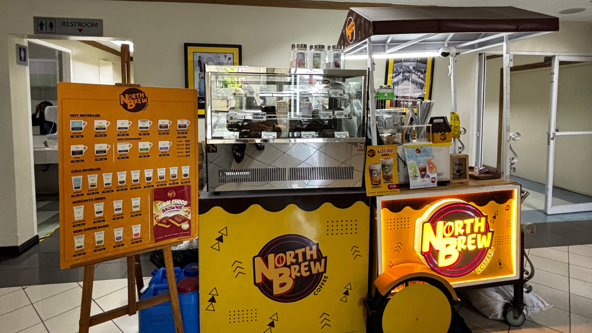 An indoor NKTI North Brew Coffee pop-up cart setup featuring a yellow illuminated logo sign, a commercial espresso machine, and a standing wooden menu board.