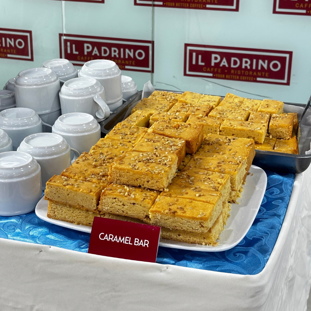 Stacks of square caramel bars topped with crushed nuts served on a dessert platter next to white coffee cups.