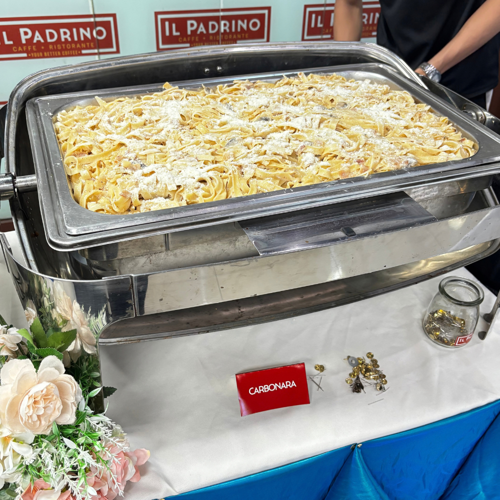 A large stainless steel chafing dish filled with creamy carbonara pasta and topped with grated cheese.