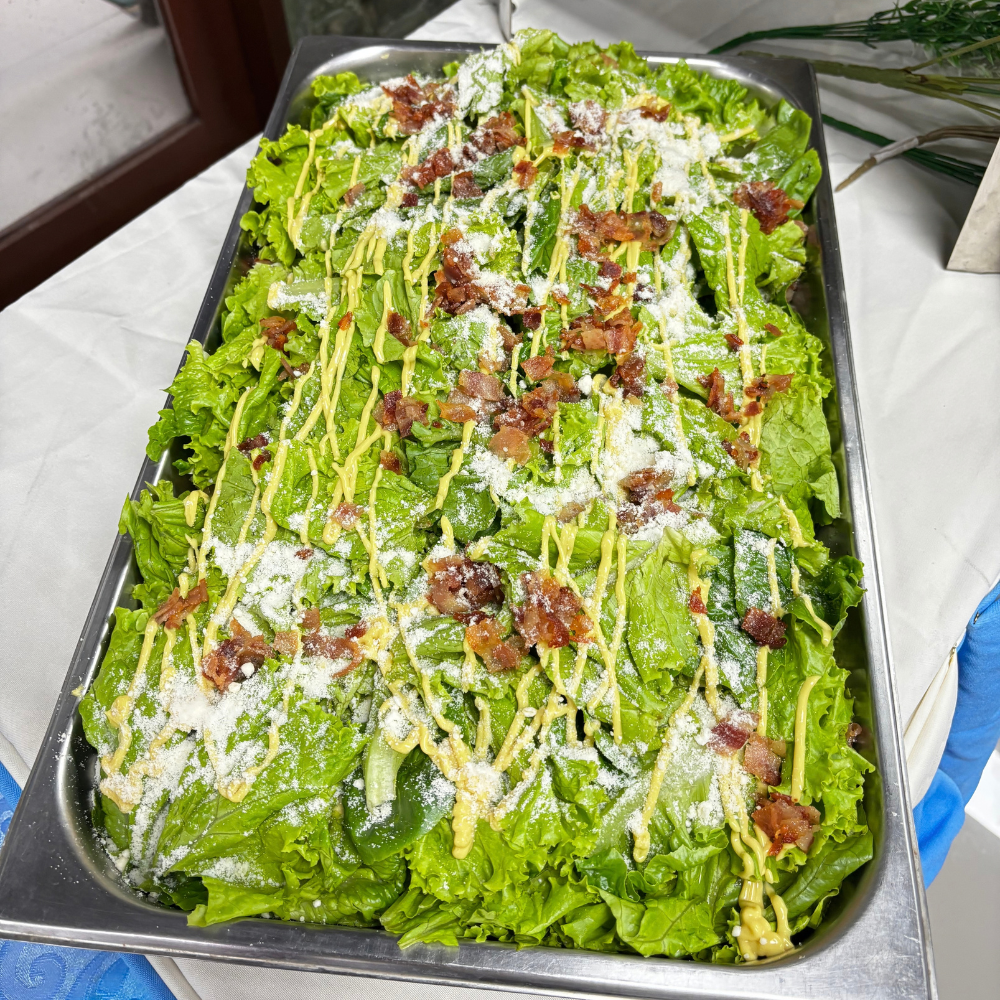 A large rectangular catering tray filled with fresh Caesar salad, topped with dressing, grated cheese, and bacon bits.