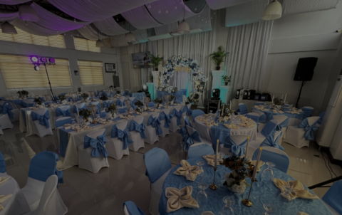 An elegant banquet hall setup for a reception at Il Padrino Events Place, displaying round tables covered in blue linens and white chairs tied with blue ribbons.