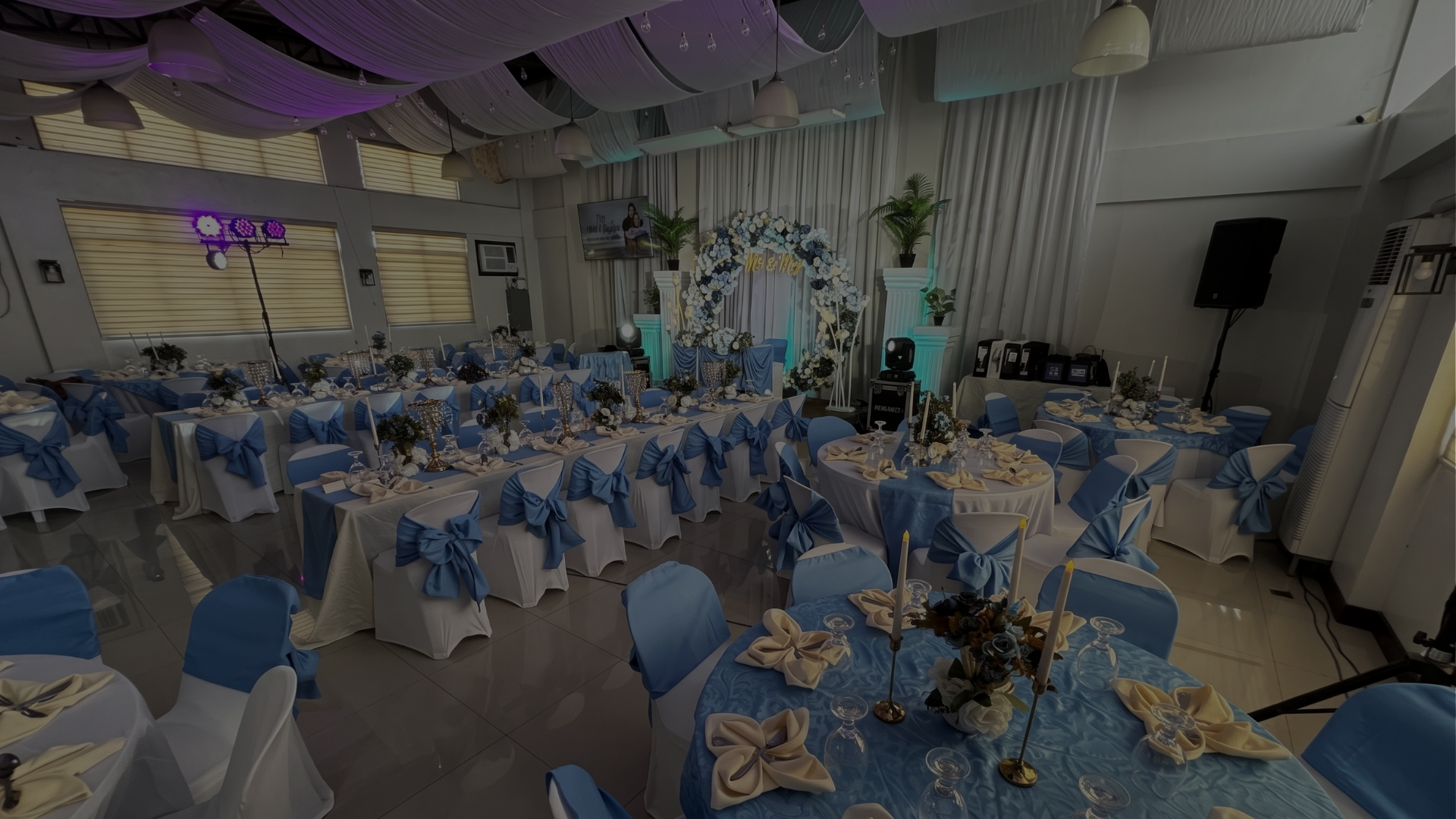 An elegant banquet hall setup for a reception at Il Padrino Events Place, displaying round tables covered in blue linens and white chairs tied with blue ribbons.
