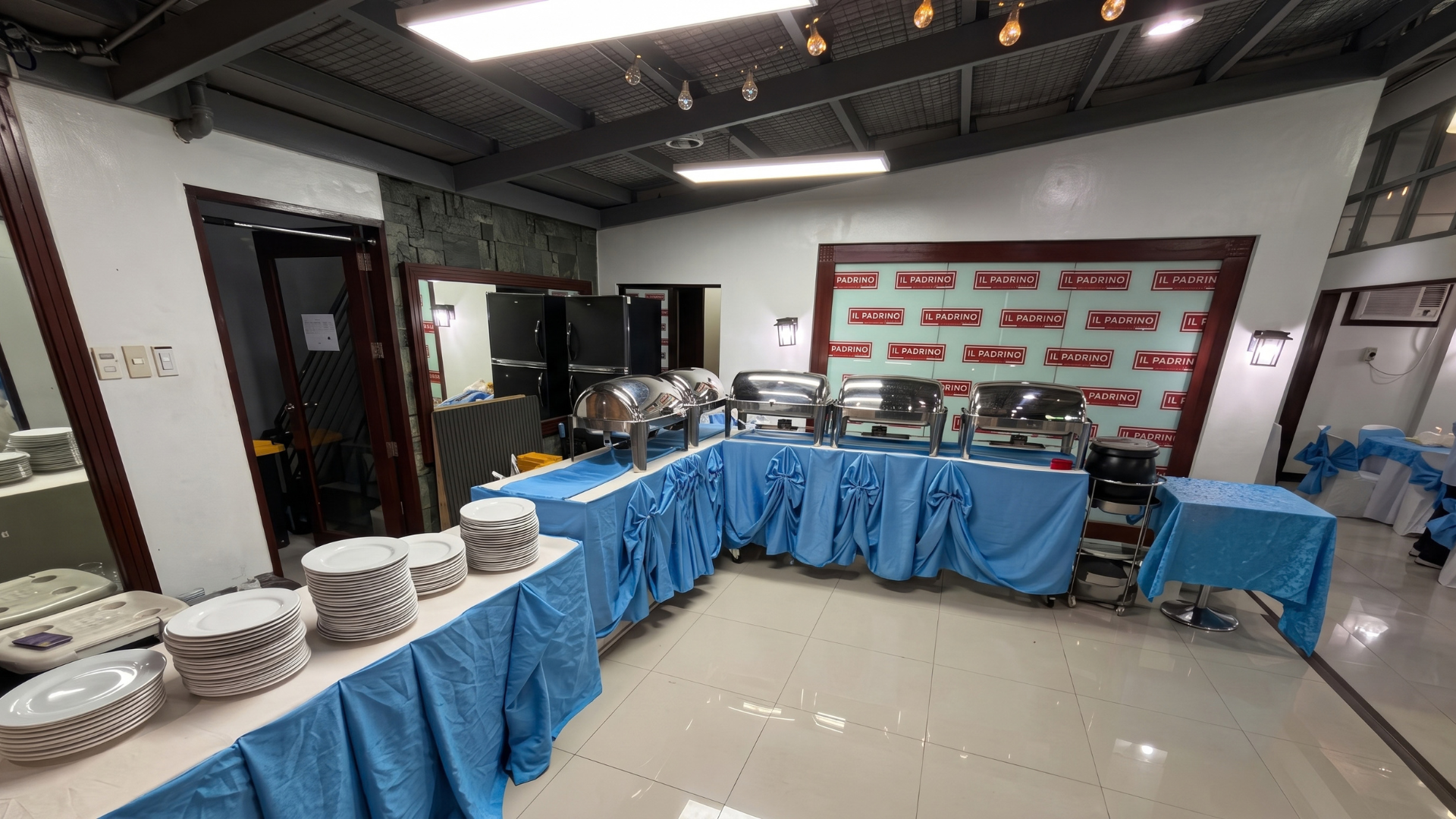 A buffet catering setup at Il Padrino Events Place, featuring stainless steel chafing dishes and stacks of white plates on a table with blue skirting.