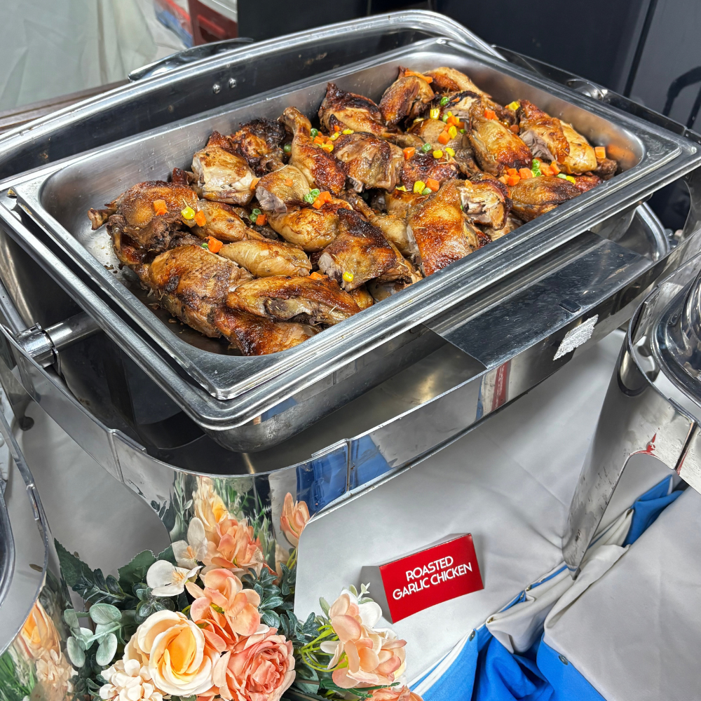 A chafing dish serving roasted garlic chicken garnished with mixed vegetables on a buffet table.